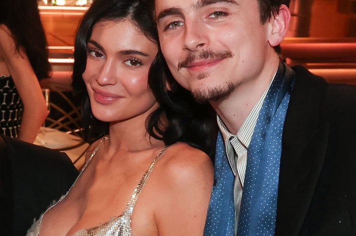 A smiling man and woman pose at a glamorous event. The woman wears an elegant, shimmering dress, while the man is dressed in a suit with a scarf
