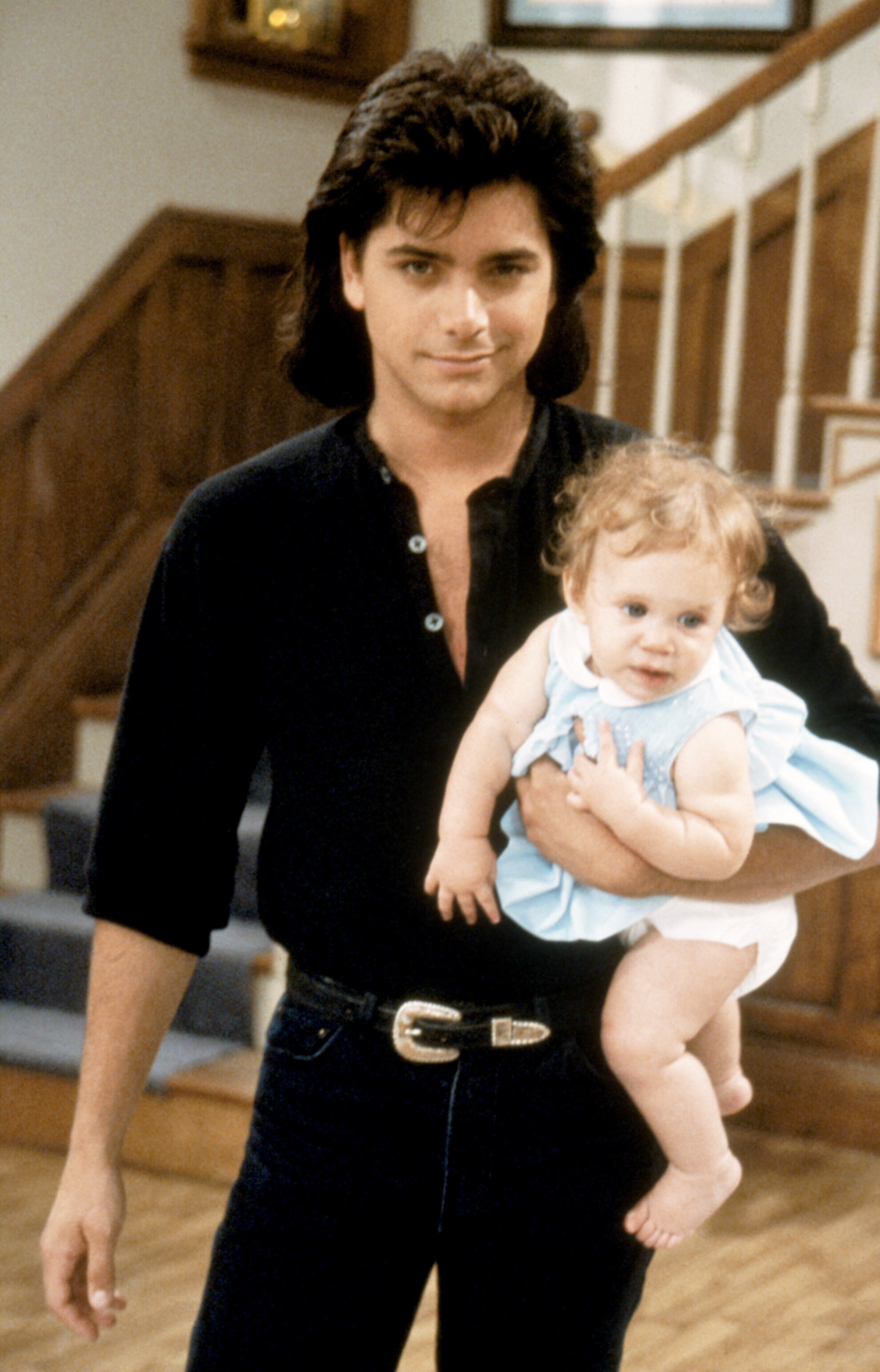Person holding a baby on a TV show set. They're wearing casual 80s-style clothes with a button-up shirt and jeans