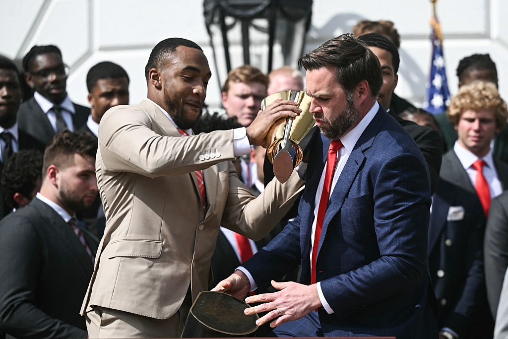 Two men on a stage, one in a beige suit pours something from a trophy over another man in a blue suit. A group of people in suits watch
