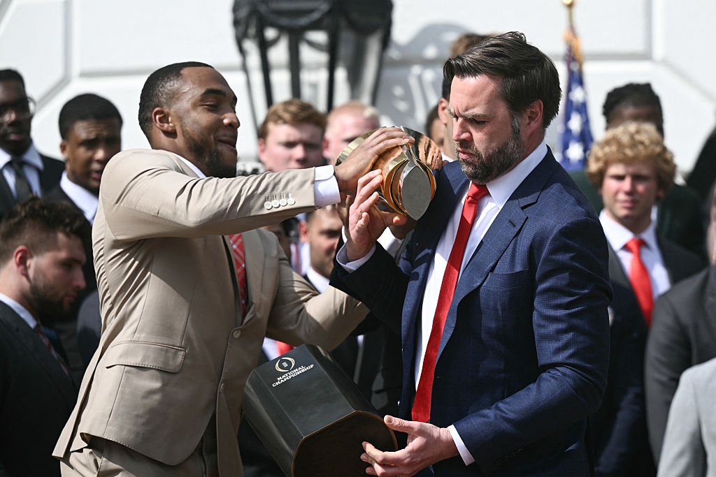 A man playfully pours a drink on another man in a suit holding a trophy, in a group setting