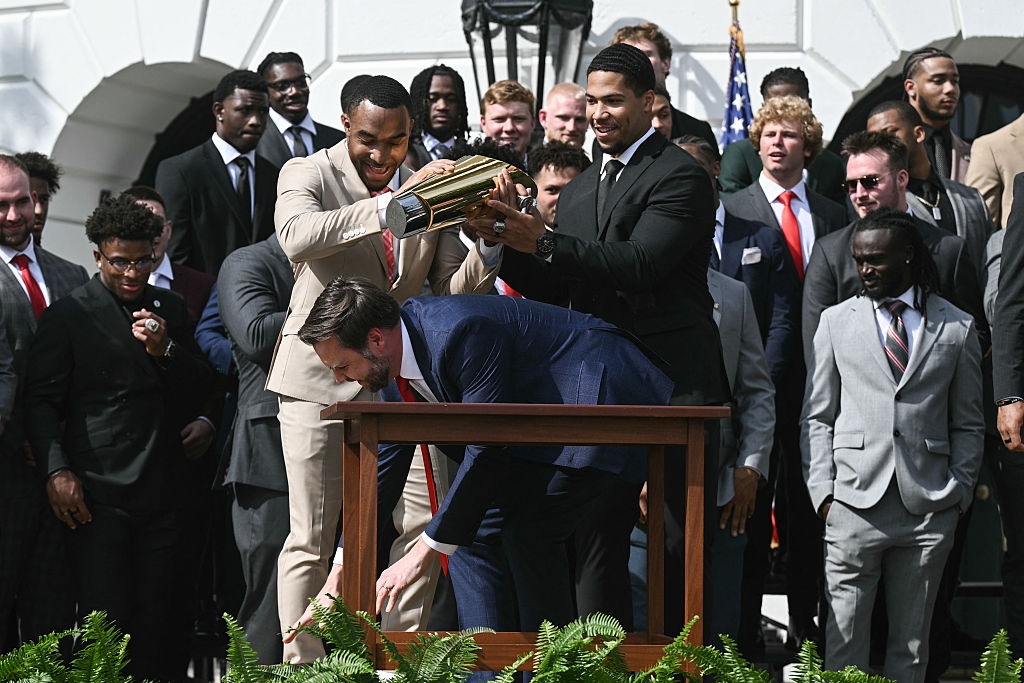 Group of suited men in a playful scene, simulating a trophy presentation, gathered on a stage with a large audience behind them