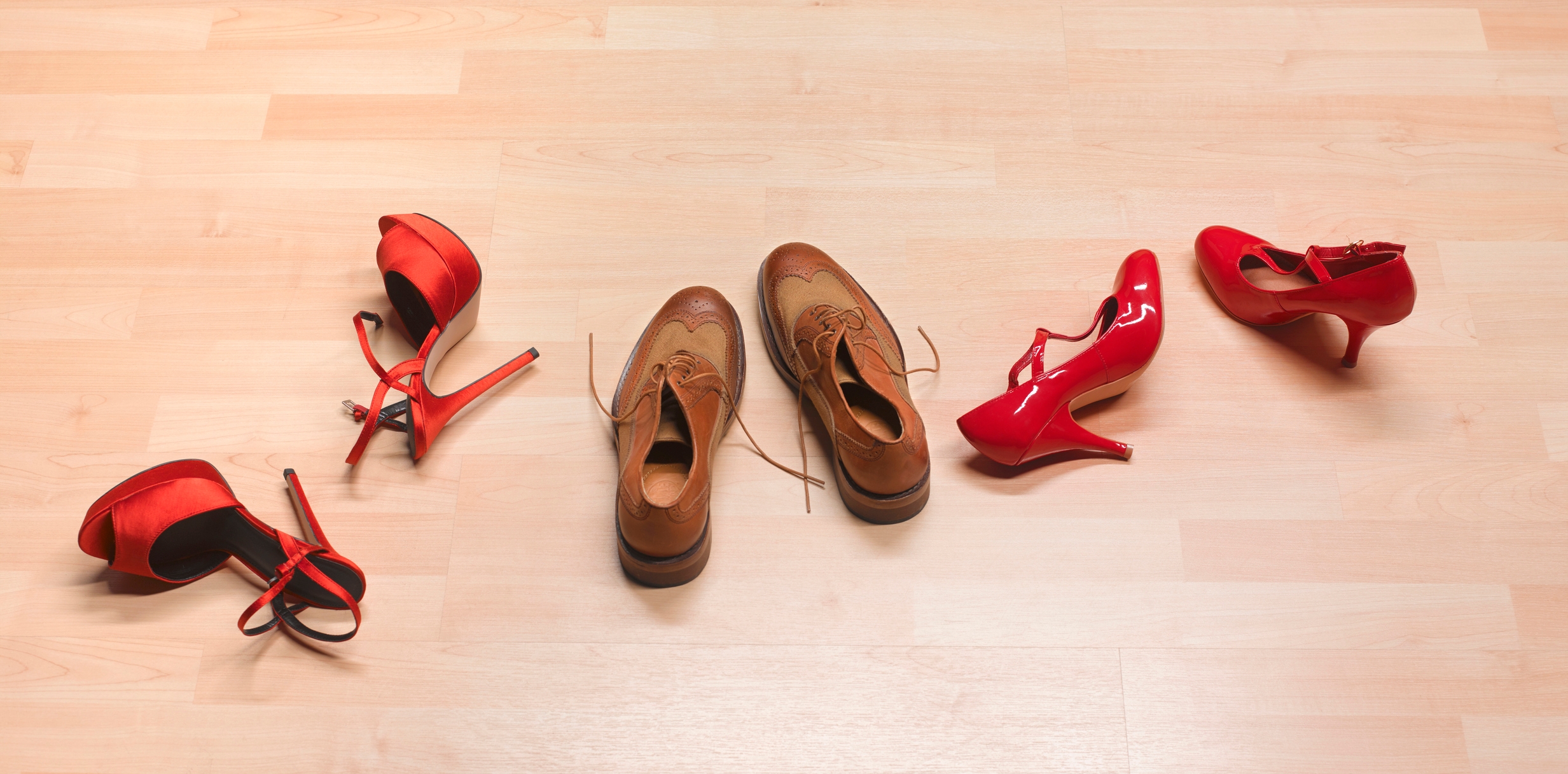 Two pairs of red high heels and a pair of brown dress shoes arranged on a wooden floor, suggesting a romantic or playful theme