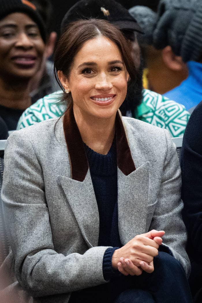 Meghan Markle smiling, seated, wearing a tailored blazer with a dark collar and a sweater underneath. Background shows other people seated closely