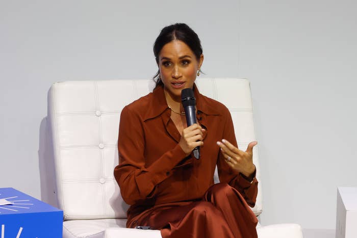 A woman sits on a stage holding a microphone, wearing a long-sleeved, satin outfit. She is speaking and gesturing with her other hand