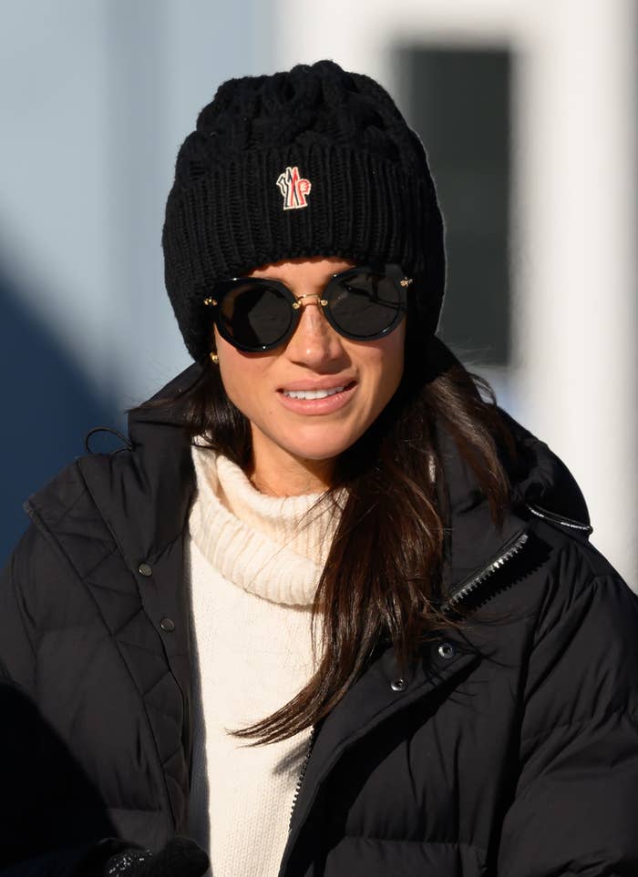 Meghan Markle wearing a knit beanie, sunglasses, white sweater, and puffy jacket outdoors