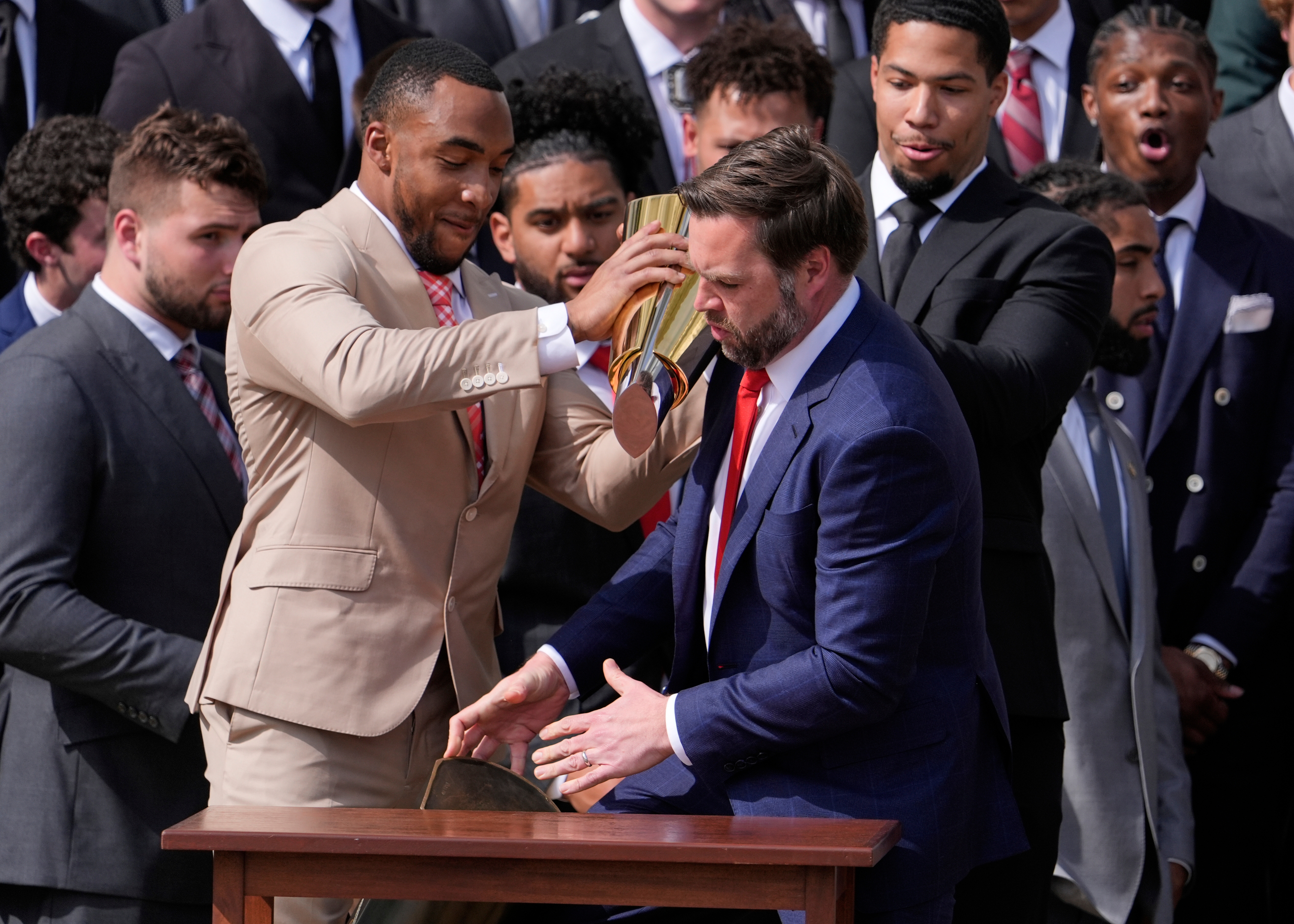 Two men in suits share a playful moment during a formal event. One pours something from a trophy over the other, while a group looks on with amusement