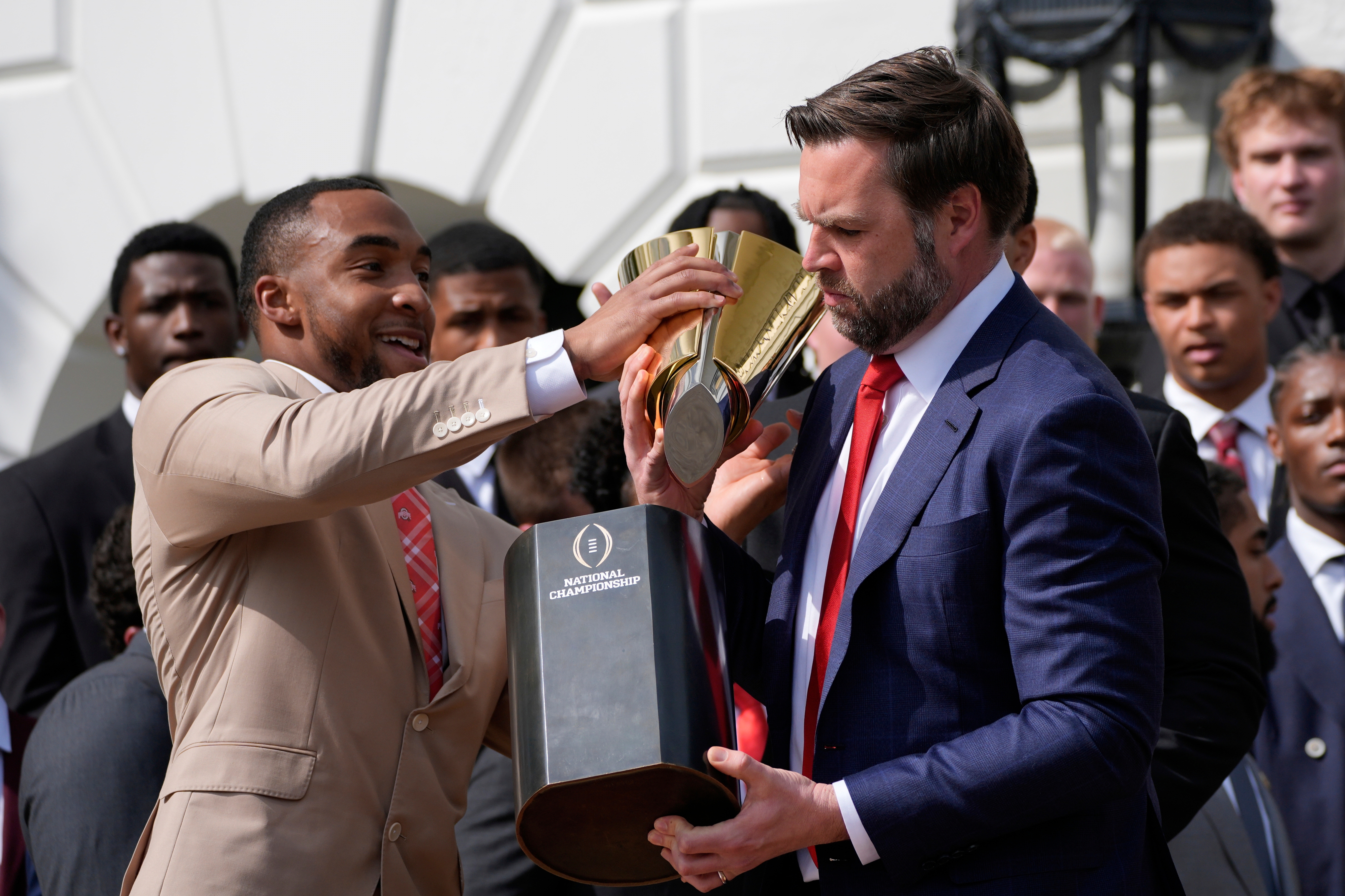 Group celebrating with football trophies; one man in a suit hands over a trophy to another. They are in a formal setting with others watching