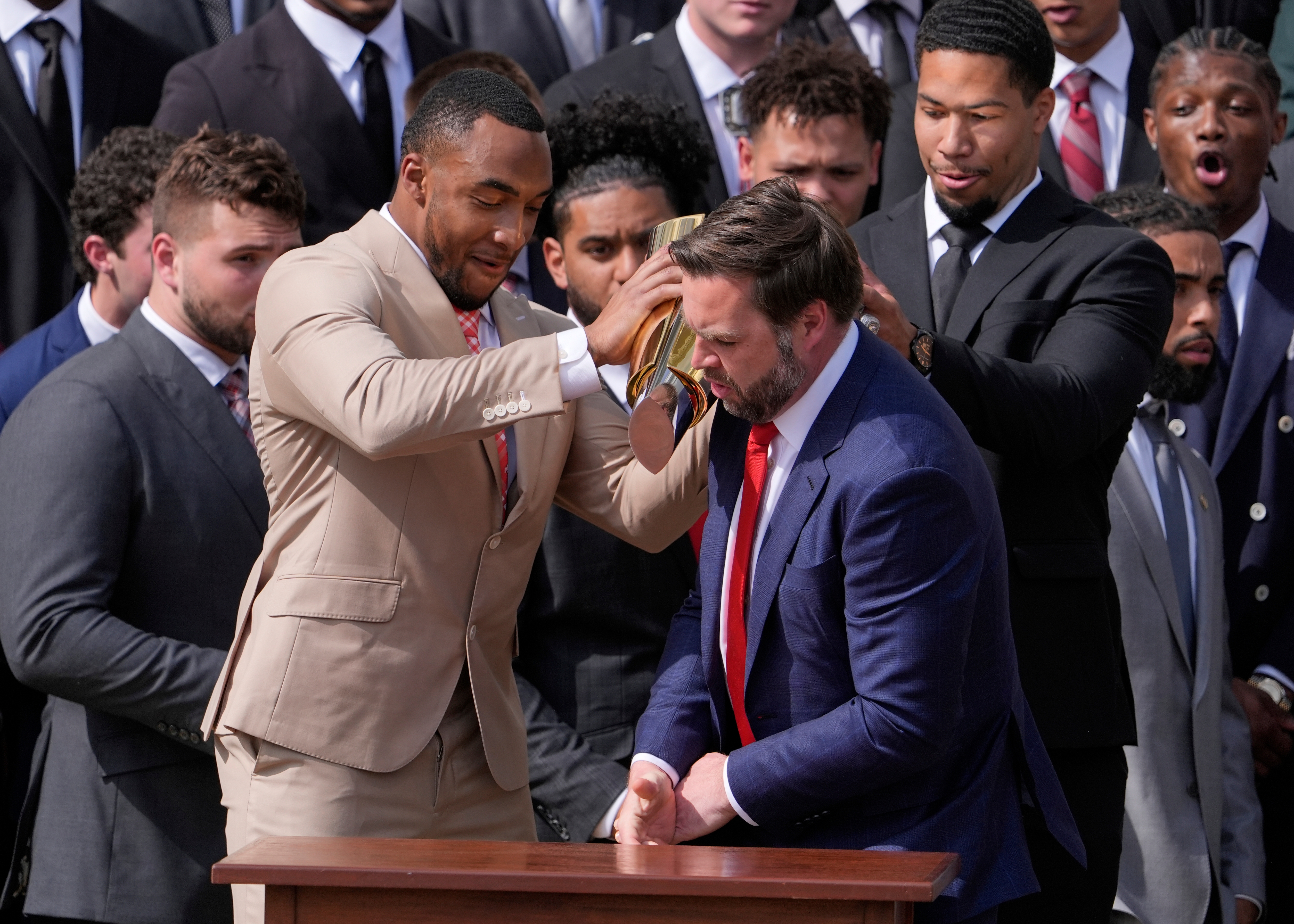 A man in a tan suit places a gold football helmet on another man's head as they are surrounded by others in suits