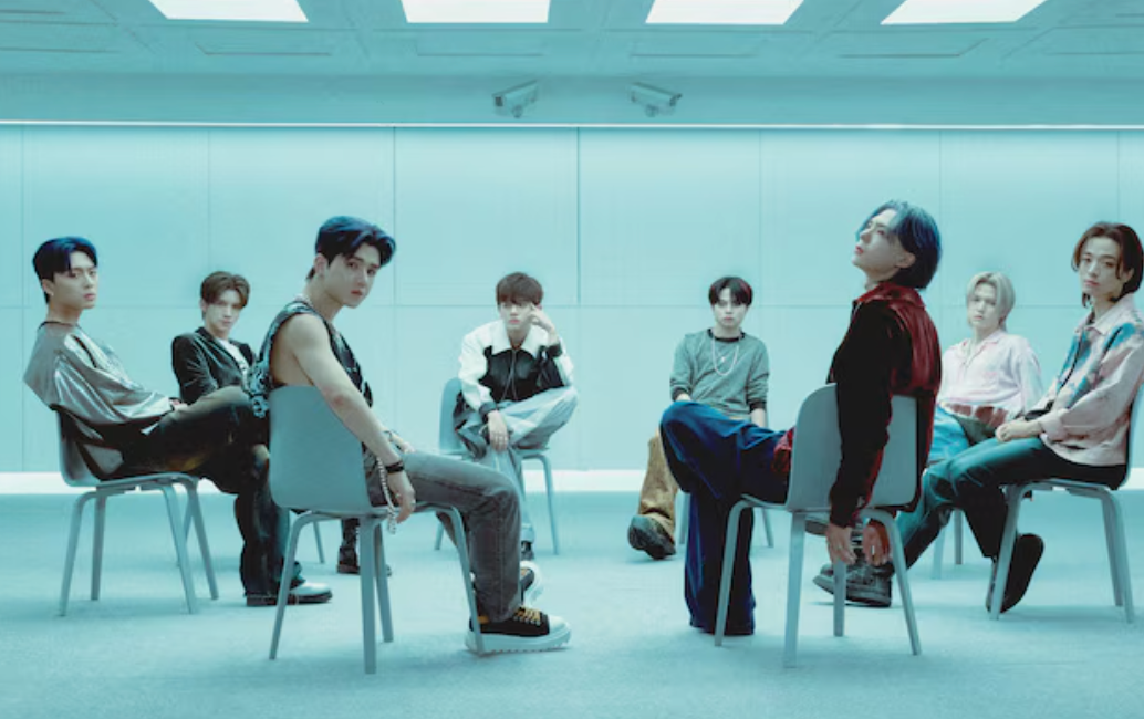 Grupo musical de ocho personas posando en sillas en una habitación moderna. Algunos miran hacia la cámara y otros hacia los lados