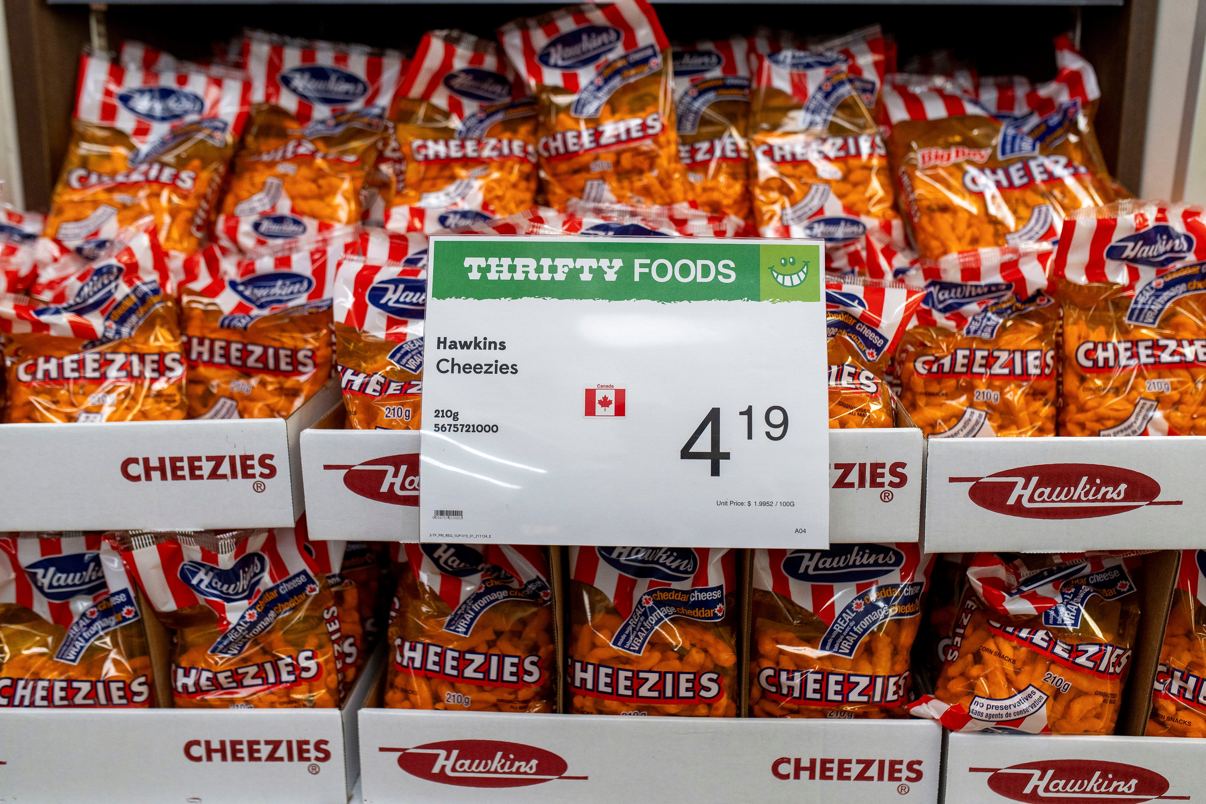 Bags of Hawkins Cheezies are displayed on shelves at Thrifty Foods with a sign showing the price as $4.19 and a Canadian flag symbol
