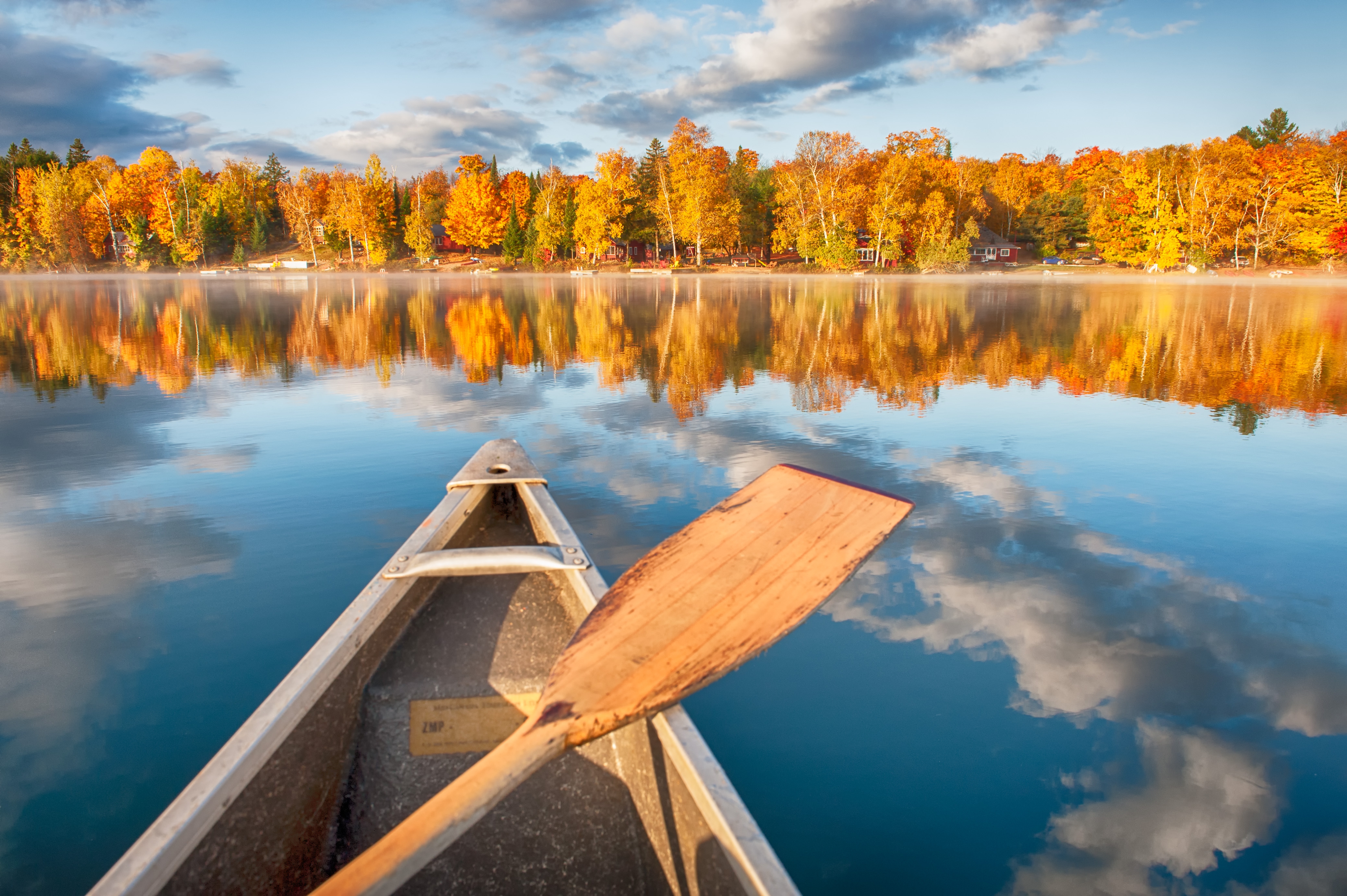 A canoe floats on a calm lake with autumn trees reflected in the water, under a partly cloudy sky