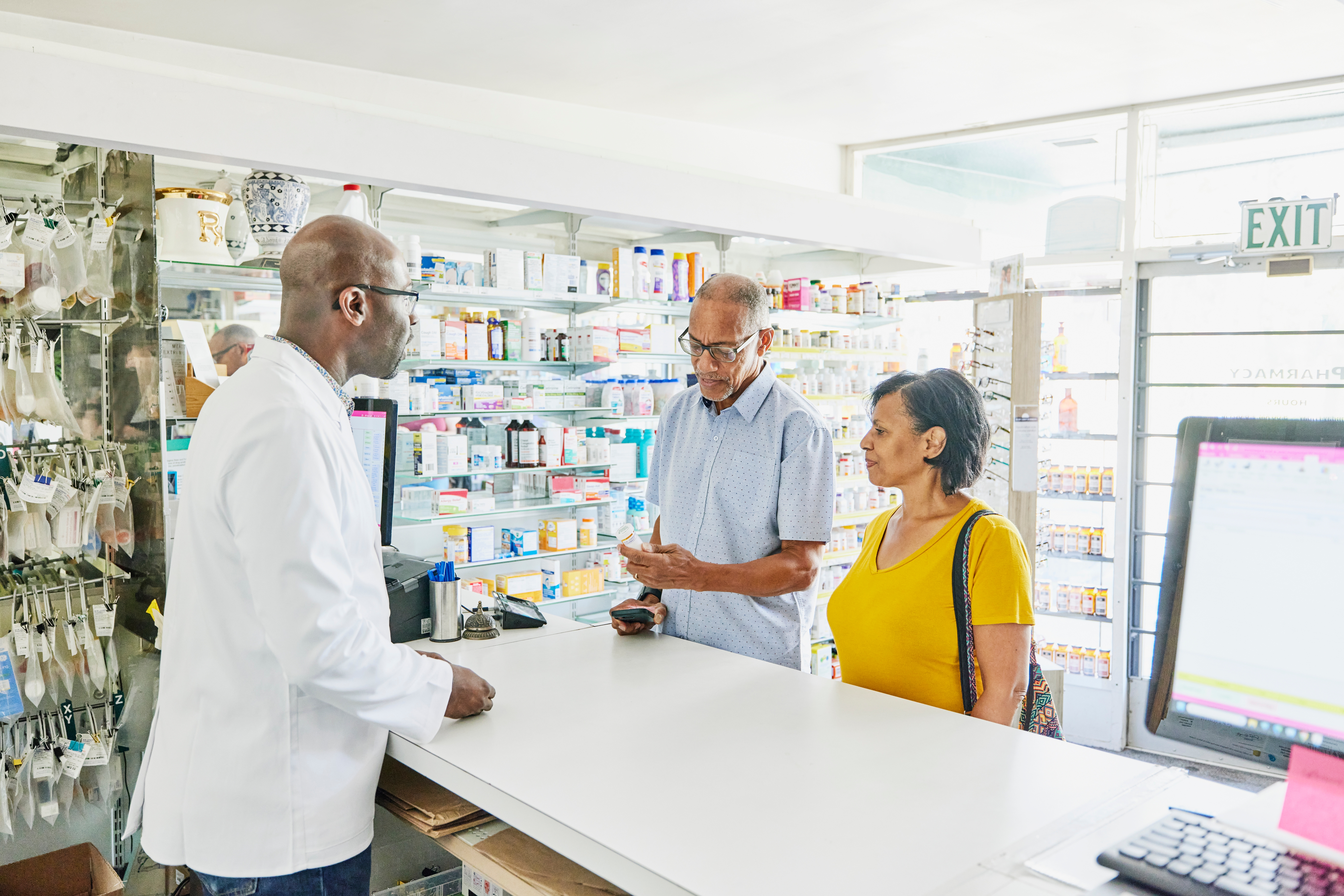 Pharmacist assists two customers at a pharmacy counter, discussing medication options. Shelves with various products in the background
