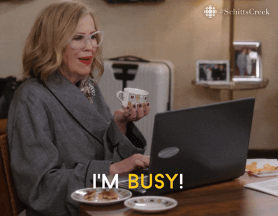 Woman in glasses smiling, holding a cup, and typing on a laptop. "I'M BUSY!" text over the image. Scene from a TV show