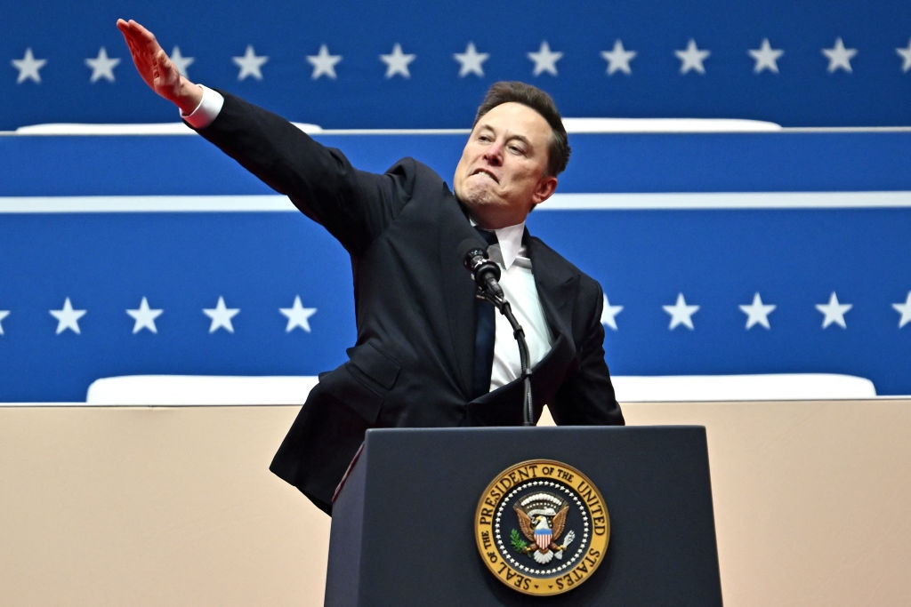 A person in a black suit makes a speech at a podium with a presidential seal, gesturing with an arm raised