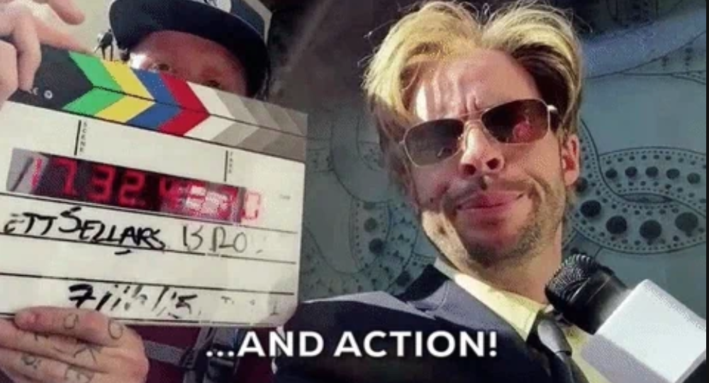A man with sunglasses stands beside a clapperboard on a film set, holding a microphone; caption reads, "…AND ACTION!"