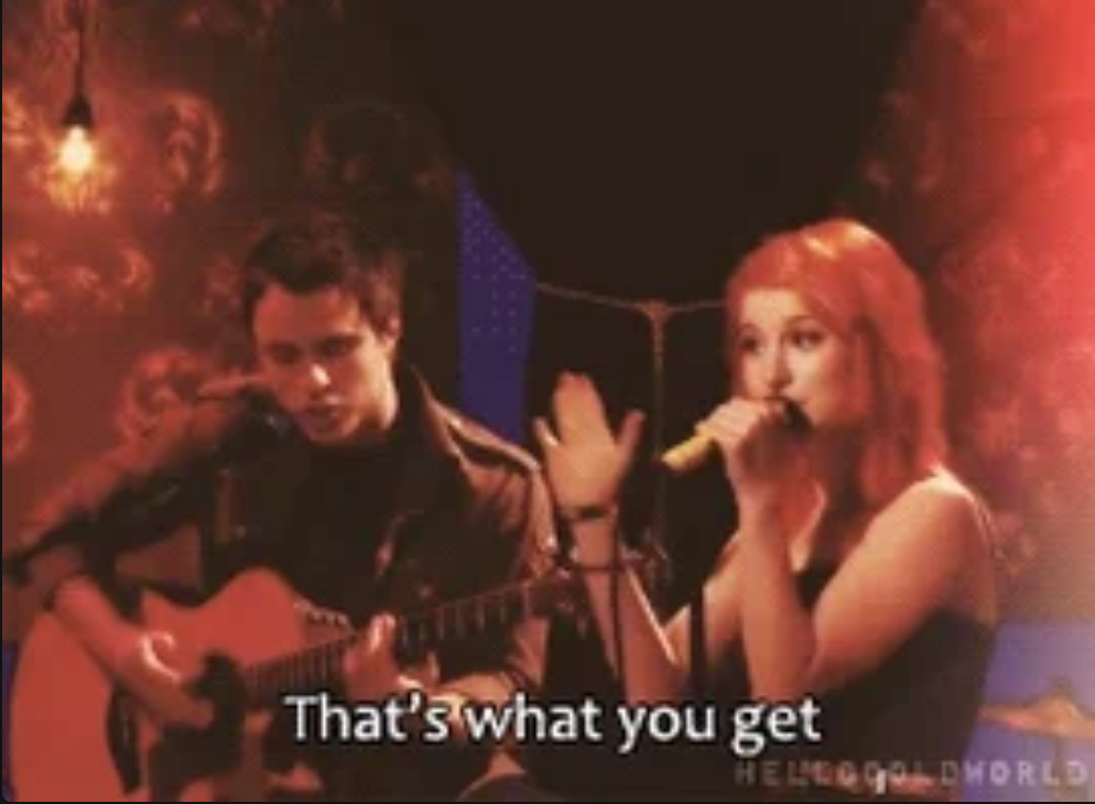 Two musicians perform on stage; the woman sings into a microphone as the man plays acoustic guitar. Subtitled: "That's what you get."