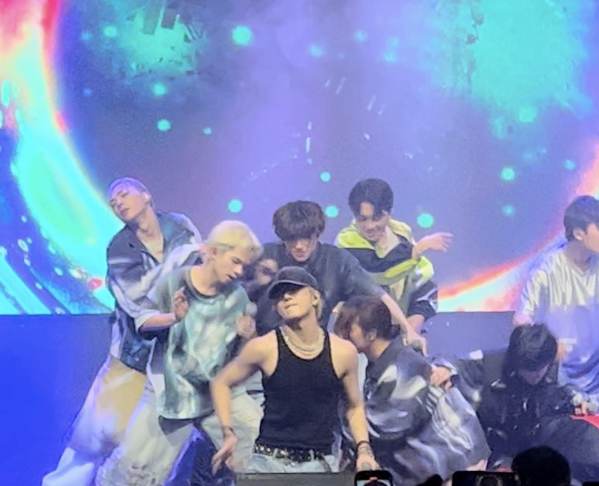 Grupo de personas en el escenario, algunas con atuendos futuristas, en medio de una actuación. Fondo con pantalla de diseño cósmico