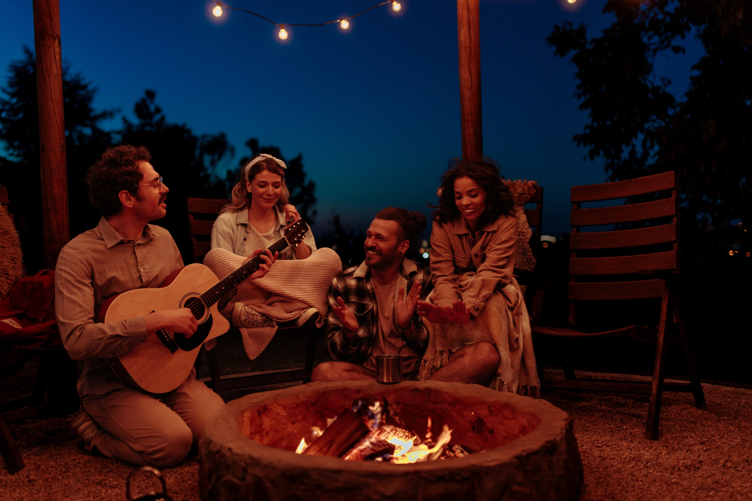 Group of friends sitting around a campfire at night. One person plays guitar, others listen and enjoy the cozy setting