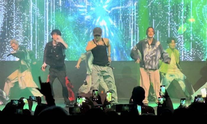 Grupo de personas baila en un concierto, rodeados de espectadores que filman con sus teléfonos. Fondo con efectos visuales brillantes