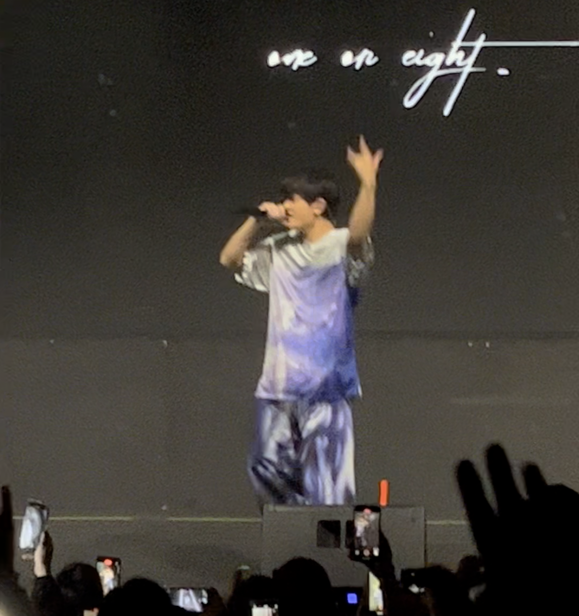 Cantante actuando en un concierto, gesticulando con las manos; público toma fotos. Texto en pared dice "on an eighth"
