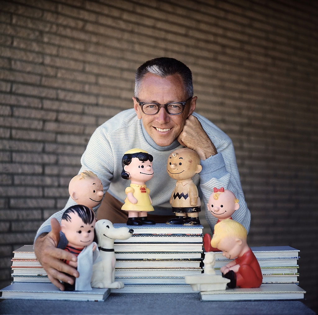 A man with glasses smiles while leaning on a table with Peanuts character figurines, including Charlie Brown and Snoopy, displayed in front of him