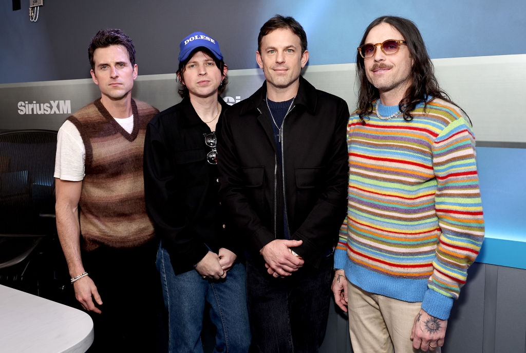 Four men stand in a studio. One wears a sweater vest, another a cap, the third a jacket, and the last a striped sweater