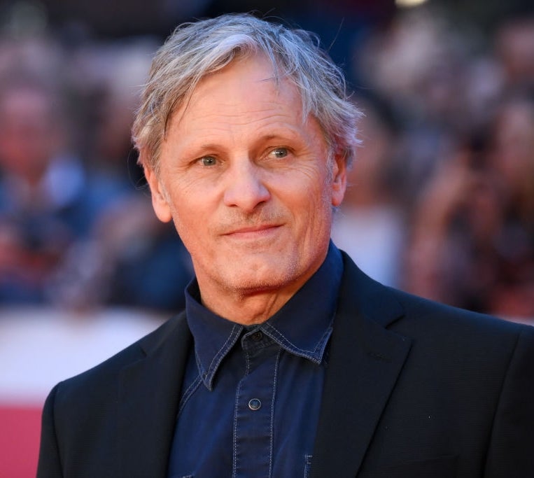 Man on red carpet in a casual dark suit with a collared shirt, looking slightly to the side. Blurred crowd in the background