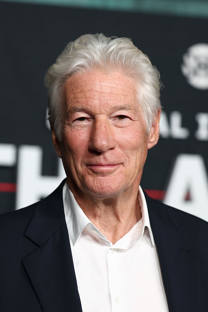 A person with white hair is wearing a suit and smiling in front of a backdrop with text