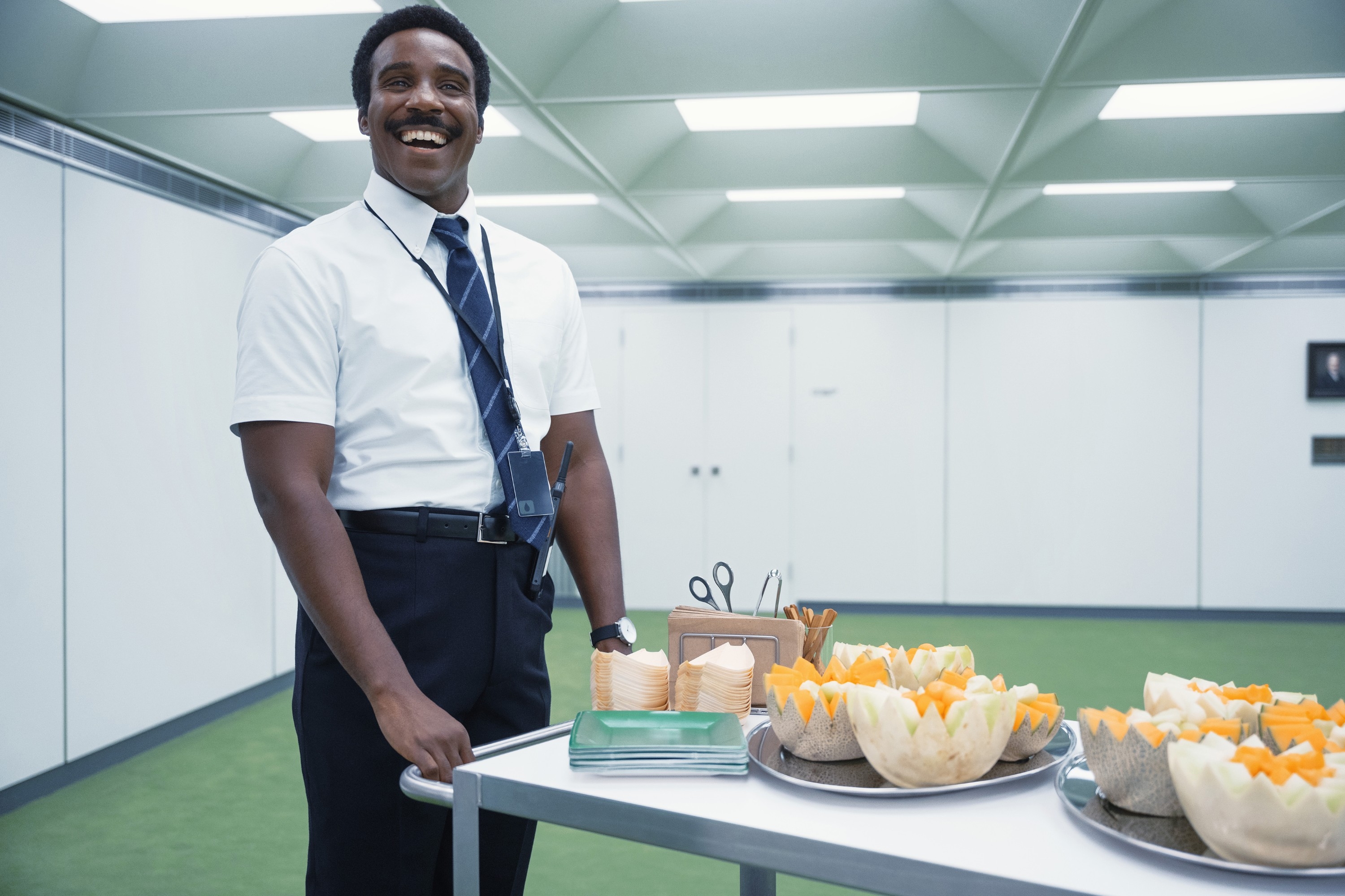 Smiling person in office attire stands beside a cart with cut fruits and napkins in a modern office setting