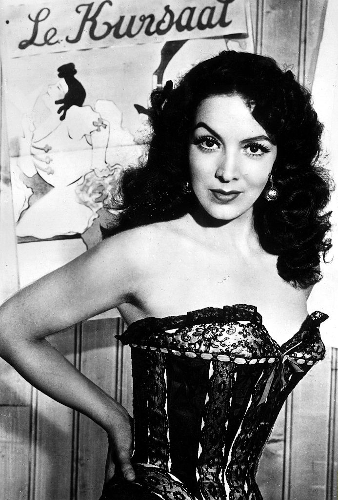 Person with vintage hairstyle in a strapless corset dress stands in front of a poster that reads &quot;Le Kursaat.&quot;