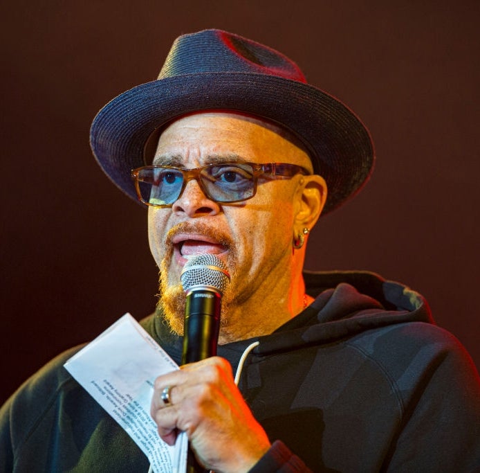 Person wearing a hat and glasses, speaking into a microphone, holding paper, on stage