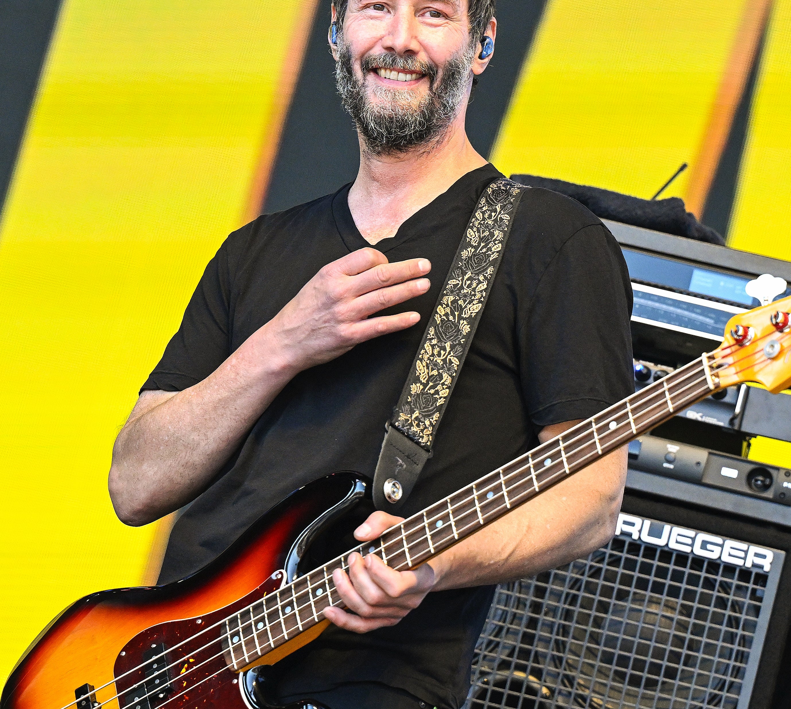 Musician on stage, smiling and holding an electric bass guitar, with amplifiers in the background