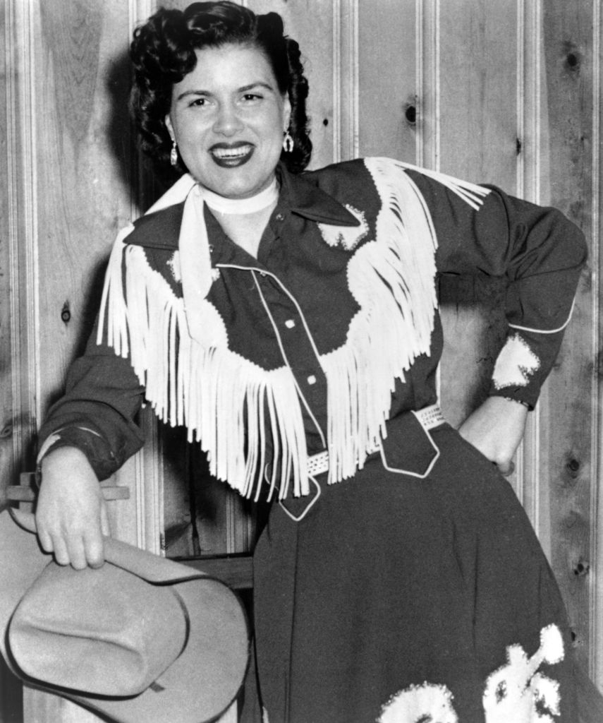 Woman in fringe cowboy outfit holding a hat, smiling in front of wood paneling