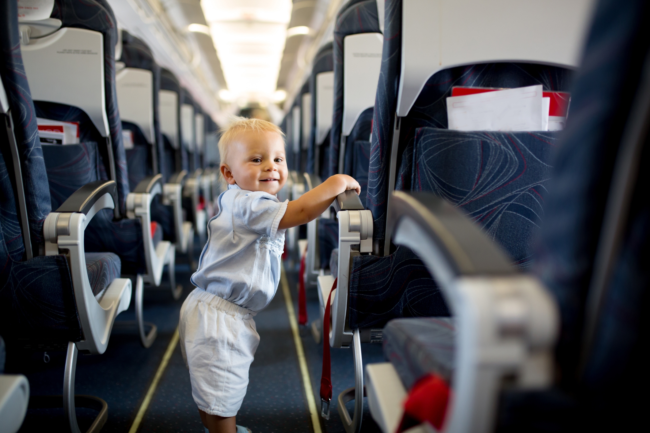 A smiling baby stands in the aisle of an airplane, holding onto a seat for balance, suggesting a family travel setting