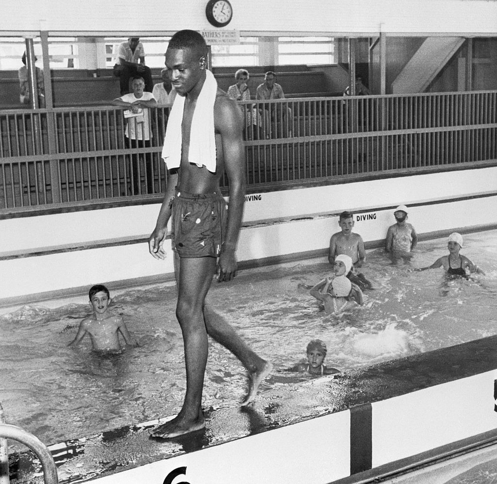 Man with towel walking by pool where kids in swim caps play; indoor setting with diving signs visible