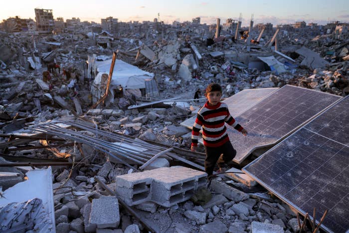 A child walks over rubble in a destroyed urban area, surrounded by debris and fallen structures. Solar panels are visible among the ruins