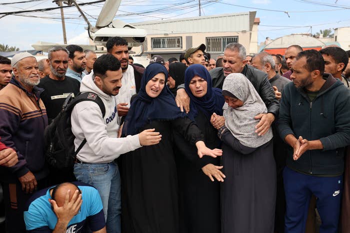 A group of grieving people is gathered outdoors, consoling each other in a crowded setting. Emotion is evident among the individuals