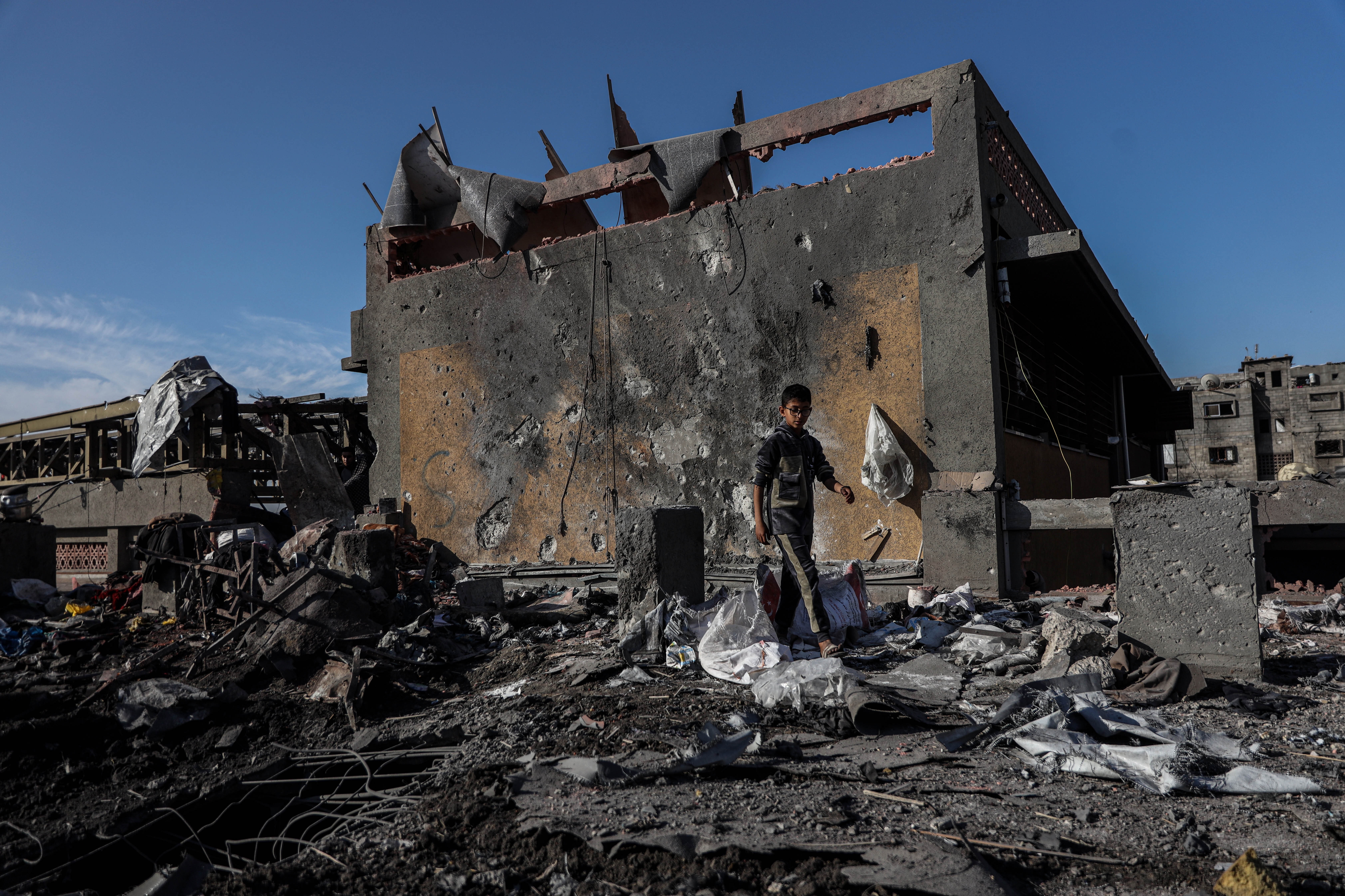 A child stands in debris before a heavily damaged, partially collapsed building in a desolate area