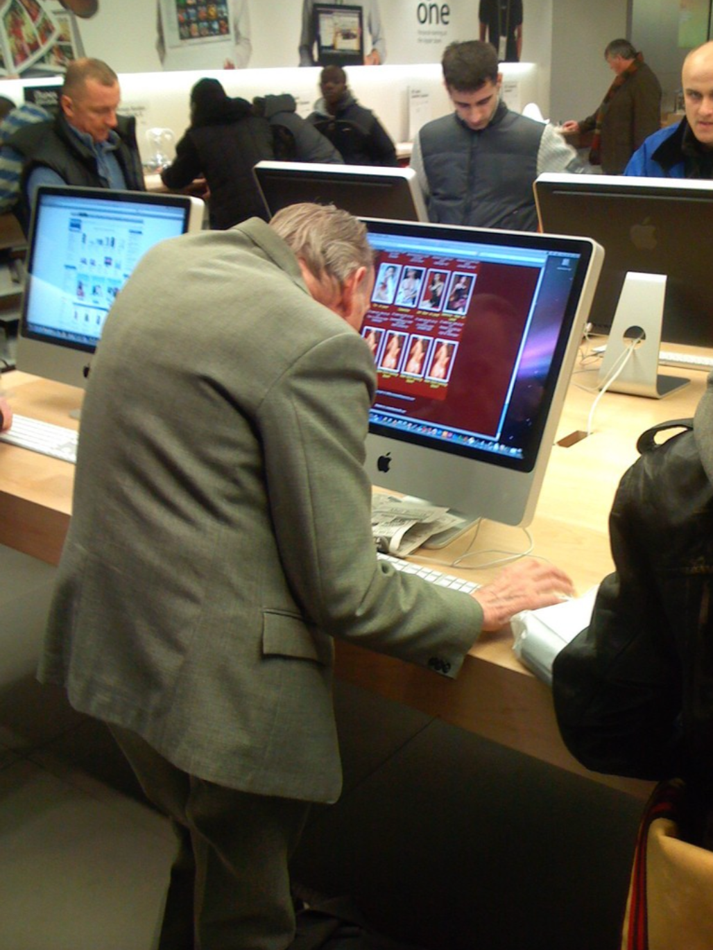 Older man examines computer screen in store, leaning in closely. Other people browse around him