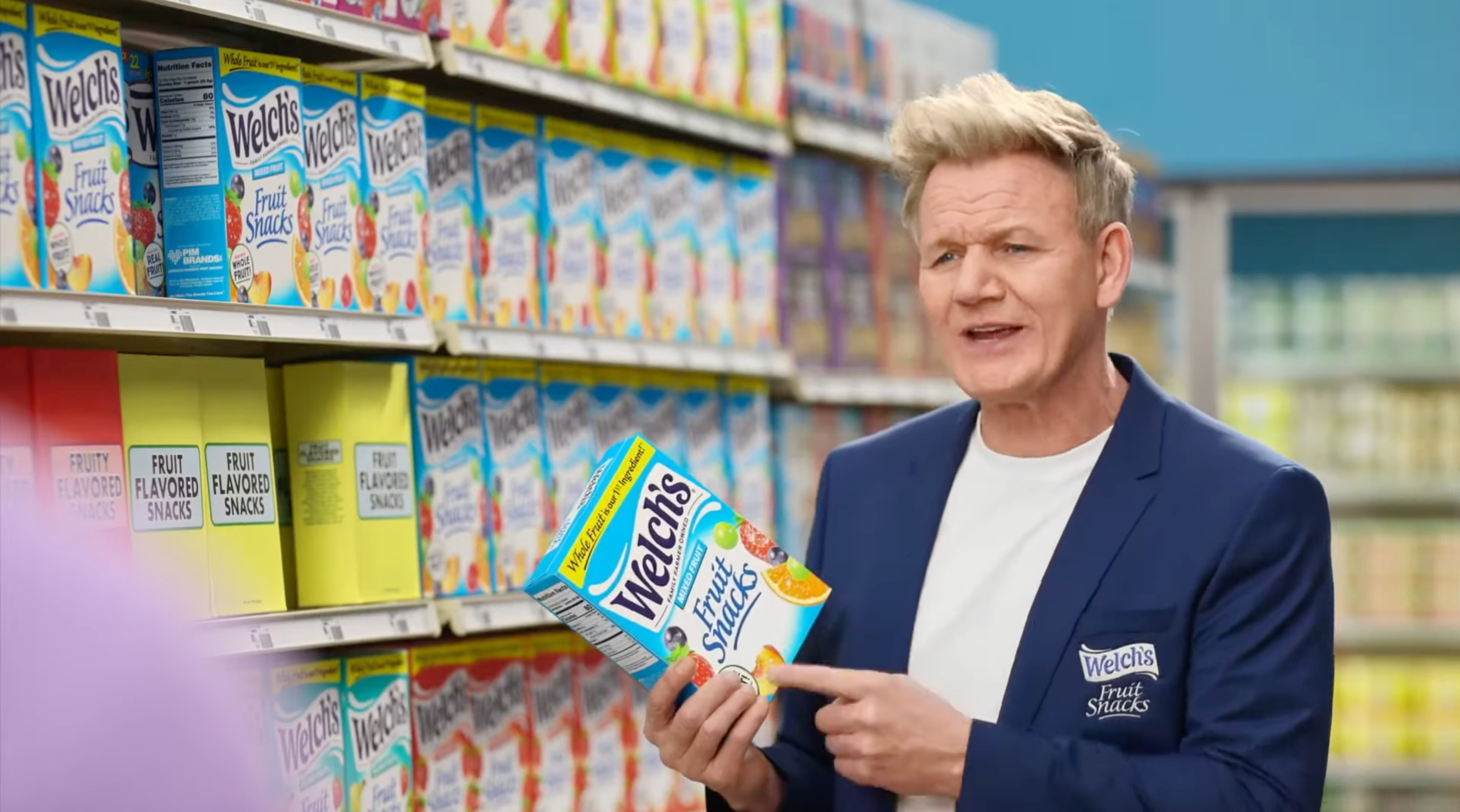 A person in a store holds a box of Welch's Fruit Snacks, pointing at it, with shelves of snacks behind them. They're wearing a blue blazer over a white shirt