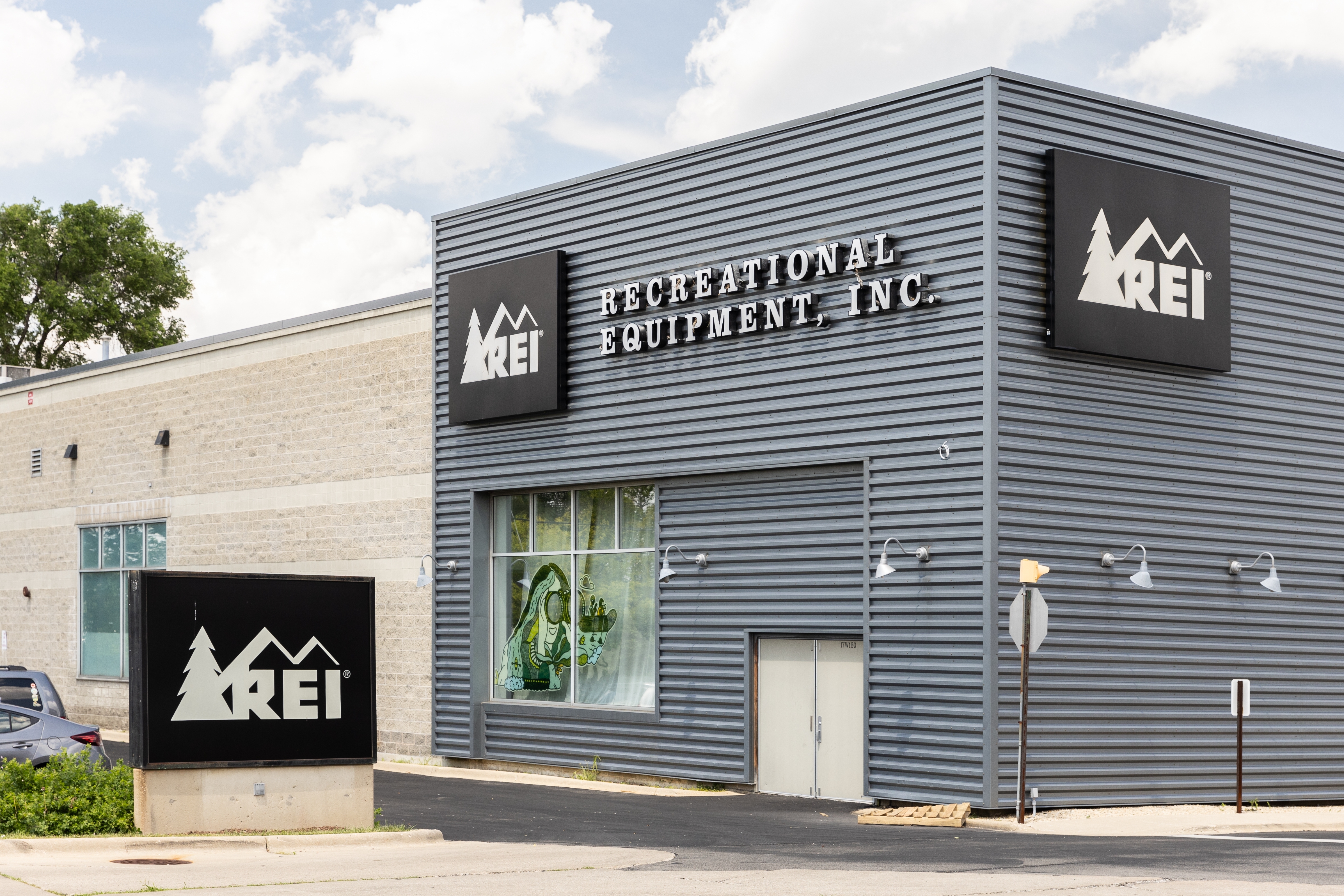 REI storefront with bold signage, showcasing outdoor gear items in window displays. Building features distinctive panel exterior