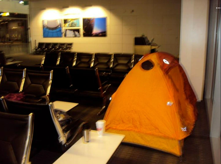 A small tent is set up in an airport waiting area next to empty seats and tables with coffee cups