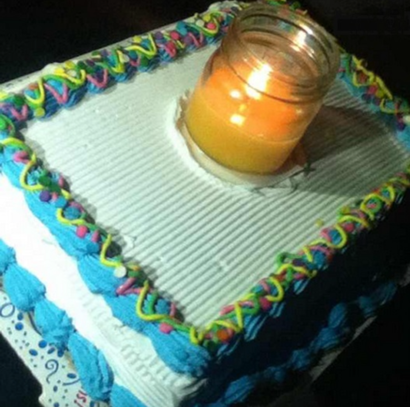 Square cake with decorative icing and a lit candle in a jar on top