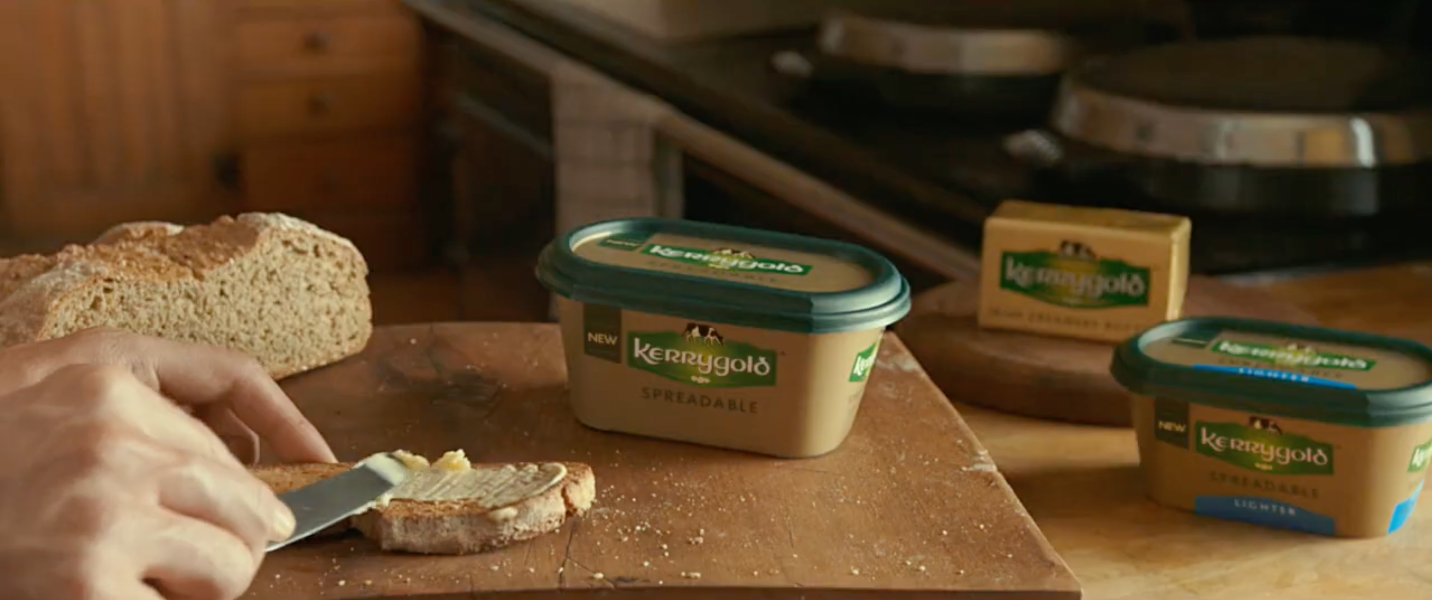 Person spreads Kerrygold butter on bread, with two other Kerrygold containers and a loaf visible on a wooden counter