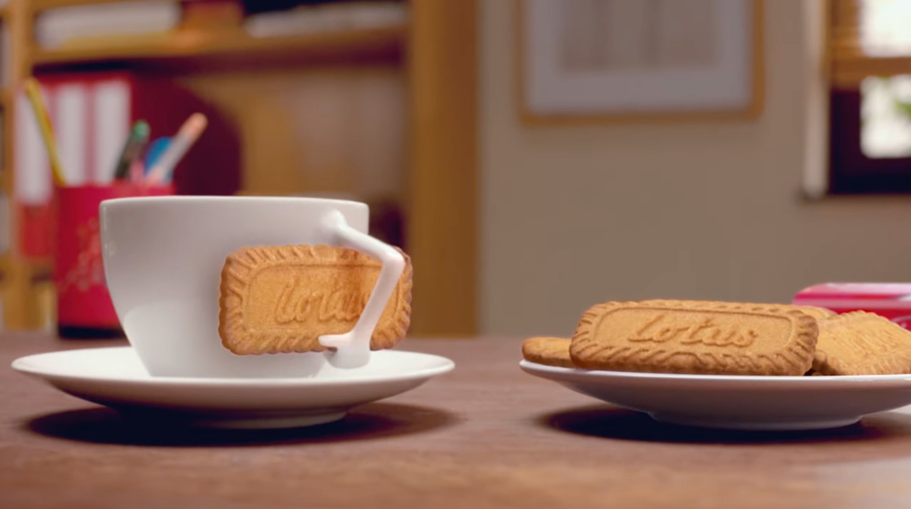 A cup with a biscuit on the saucer and a plate with similar biscuits on a table in a cozy room