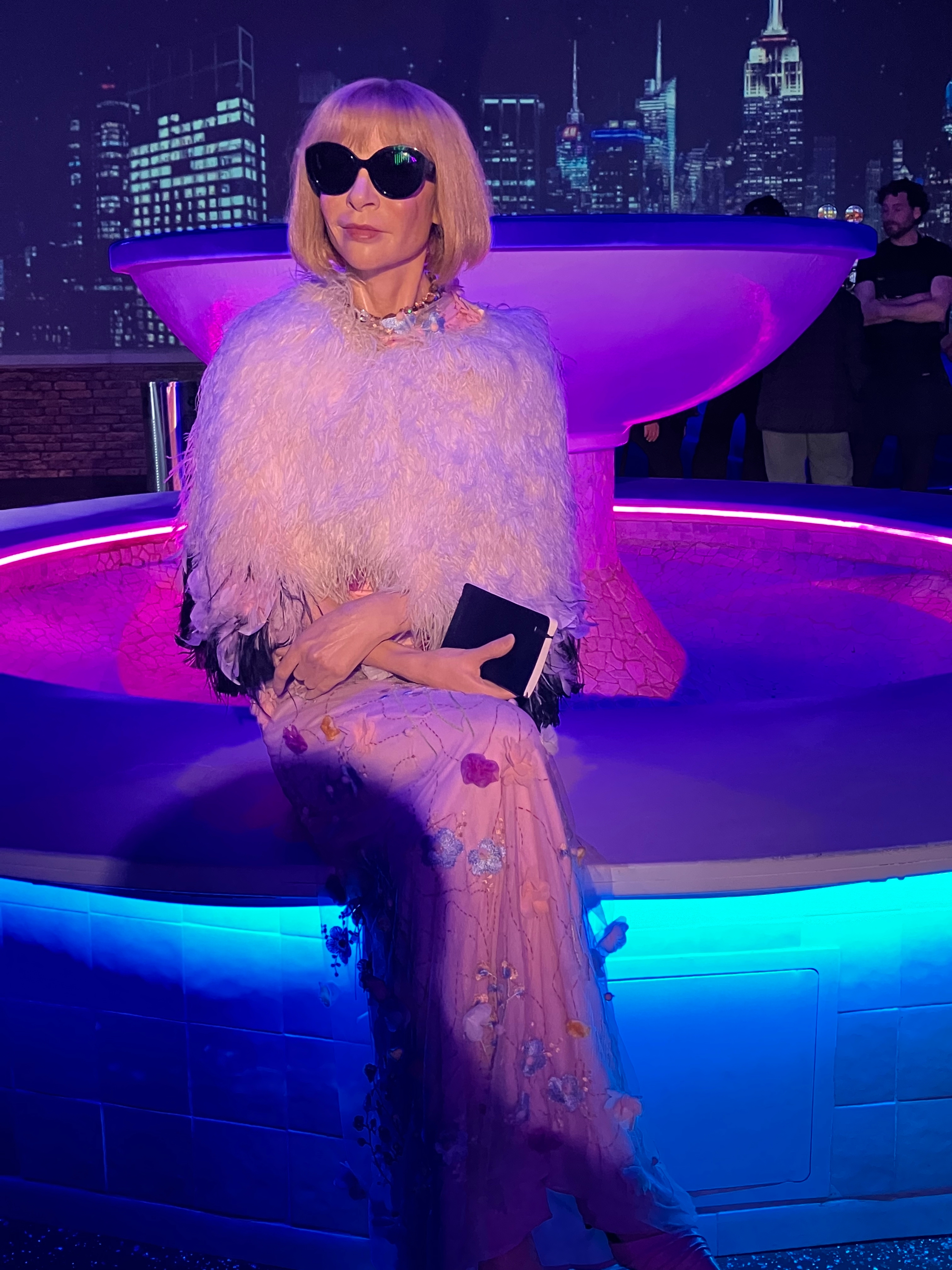 Person in a feathered wrap and embellished gown sits by an illuminated, artistic fountain with a city skyline backdrop
