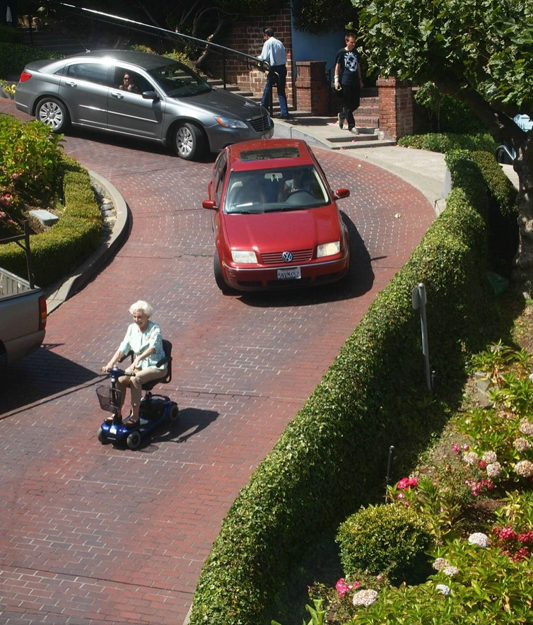 An elderly person rides a mobility scooter down a steep brick street, with a few cars following behind. Greenery lines the road's edge