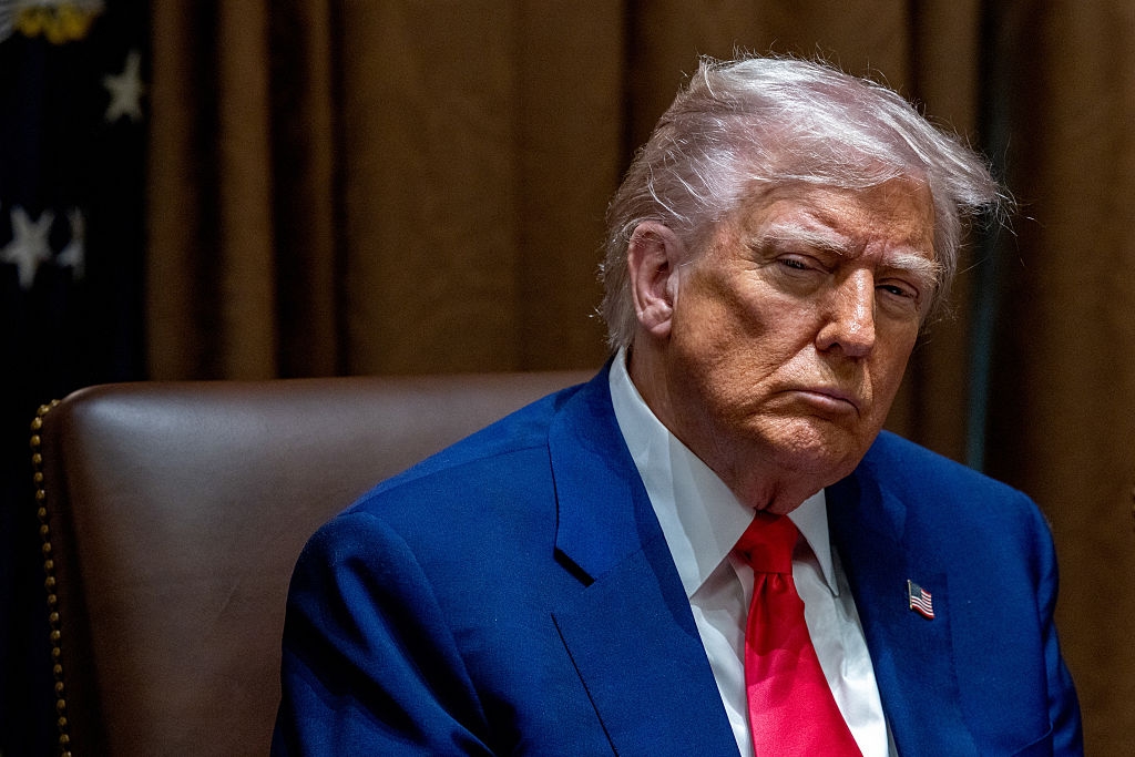 A person in a formal suit and tie sits seriously at a table in an official-looking setting