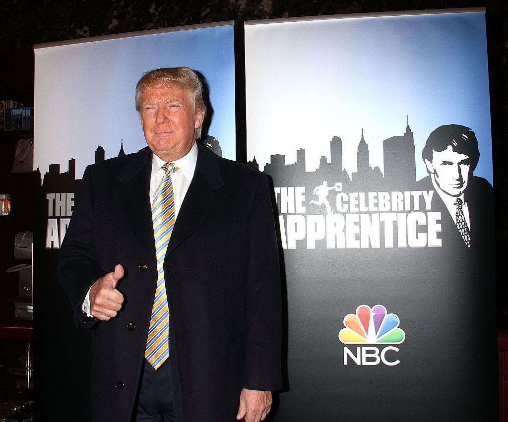 Person in a dark suit and patterned tie gives a thumbs-up in front of &quot;The Celebrity Apprentice&quot; promotional banners