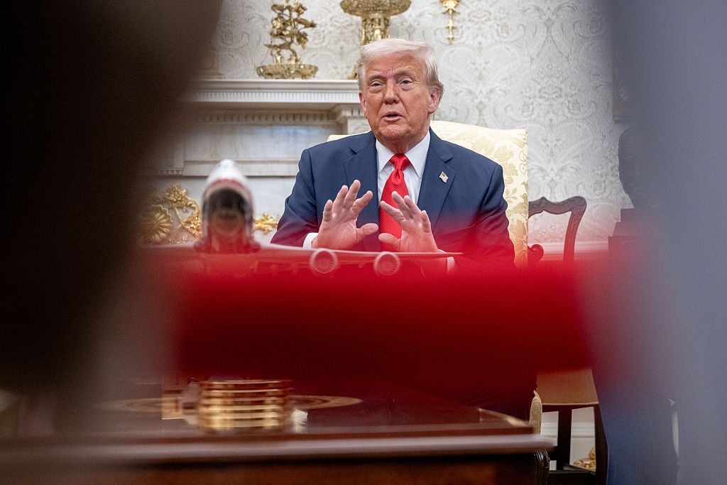 A person in a suit gestures while seated in an ornate room, with a model airplane on the table in front